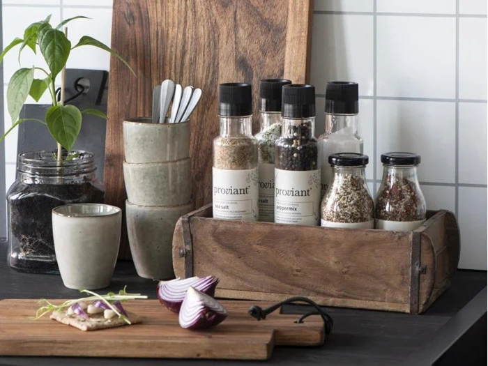 Et køkkenbord med en trækasse med krydderikrukker, stablede kopper, en plante i en krukke, træplader og hakkede rødløg på et skærebræt.