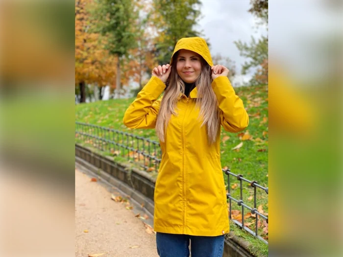 En person i en gul regnfrakke står udenfor på en sti og holder sin hætte, med efterårstræer og blade i baggrunden.