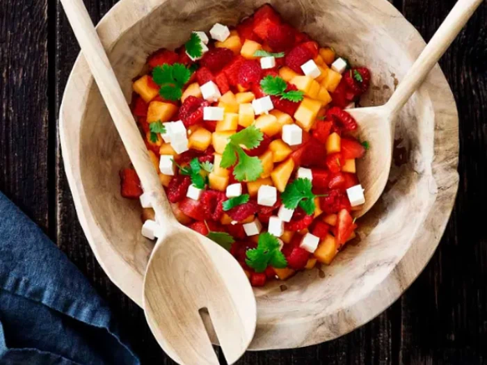 En træskål med salat lavet af jordbær, mango i tern, hvid ost og frisk koriander, med serveringsredskaber af træ indeni.