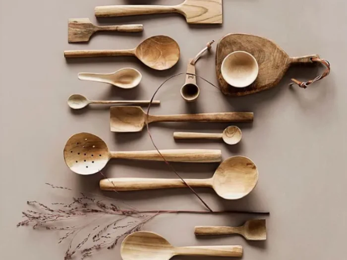 Forskellige køkkenredskaber af træ, bl.a. skeer og spatler, arrangeret pænt på en beige overflade med en tørret plantestængel i bunden.