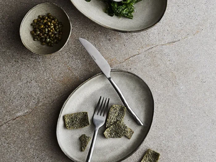 Et billede oppefra og ned af tre stentallerkener med grønne grøntsager, kapers og stykker af knækbrød samt en gaffel og kniv på en grå overflade.