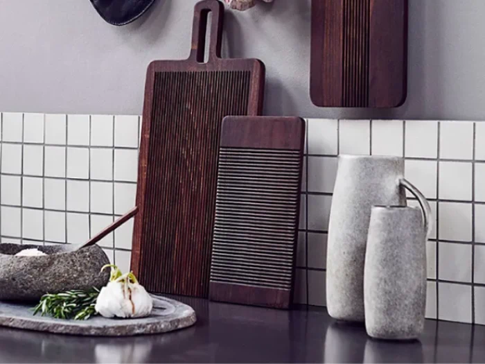 Mørke skærebrætter i træ, en stenskål med hvidløg og krydderurter og to grå keramikkander står på en mørk bordplade mod et flisebelagt stænk.