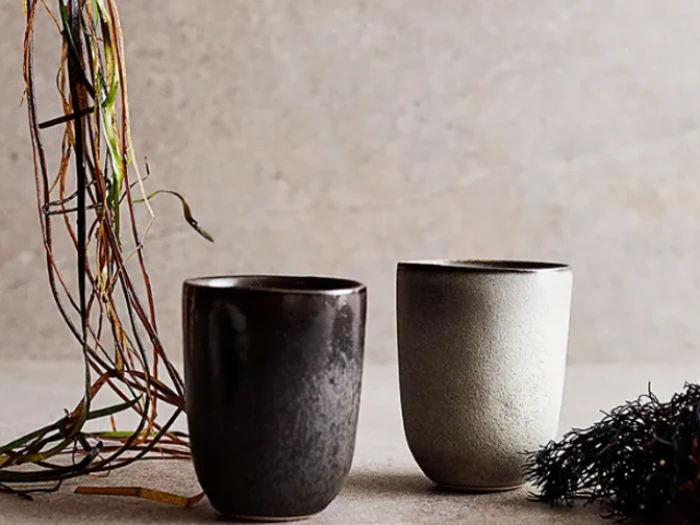 To keramiske kopper, en mørk og en lys, står på en neutral overflade med tørrede kviste og grønt ved siden af på en lys struktureret baggrund.
