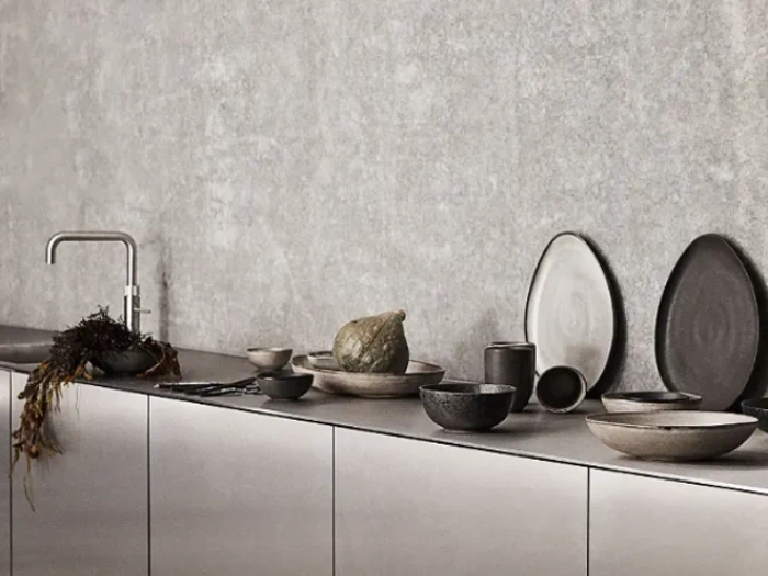 Et moderne køkkenbord med minimalistisk keramik, skåle, tallerkener, en lille squash, tang og en elegant metalarmatur mod en struktureret grå væg.