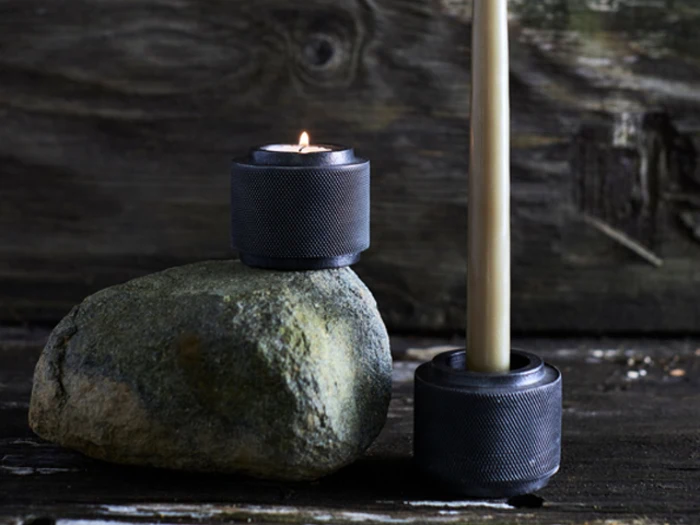 En lille sort lysestage med et tændt fyrfadslys står på en sten; ved siden af står en anden sort lysestage med et højt, utændt lys på en rustik træbaggrund.
