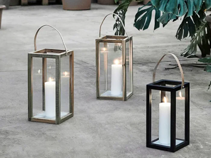 Tre glaslanterner med hvide lys indeni står på en betonflade, omgivet af grønne blade og potteplanter i baggrunden.