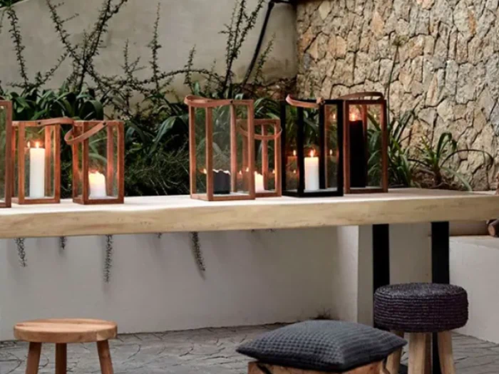 På et træbord udendørs står tændte stearinlys i glaslanterner, med skamler og puder i nærheden, mod en stenmur og grønne planter i baggrunden.