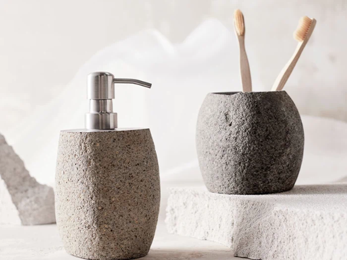 To badeværelsestilbehør med stenstruktur: en sæbedispenser og en kop med to tandbørster af træ, placeret på en hvid struktureret overflade.
