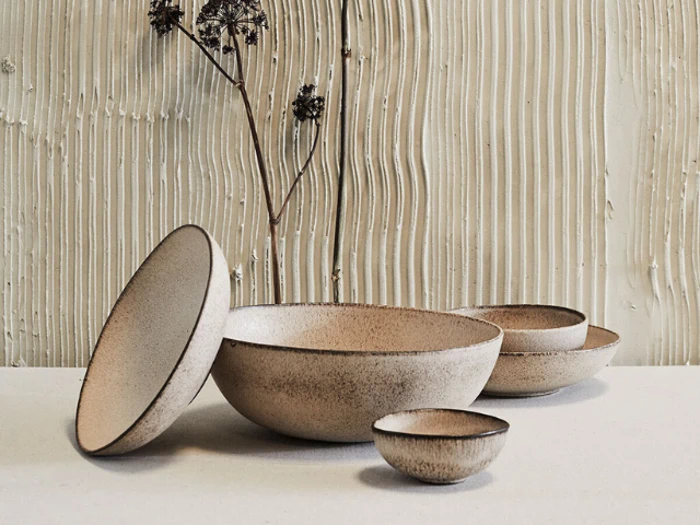 Fem keramikskåle i jordfarver er arrangeret på en lys flade, med en skål vippet oprejst; tekstureret væg og tørret plante i baggrunden.
