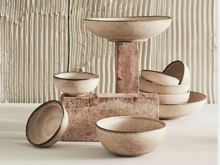 Et sæt beige keramikskåle med mørke kanter, kunstfærdigt arrangeret på og omkring strukturerede mursten mod en ribbet, neutraltonet baggrund.