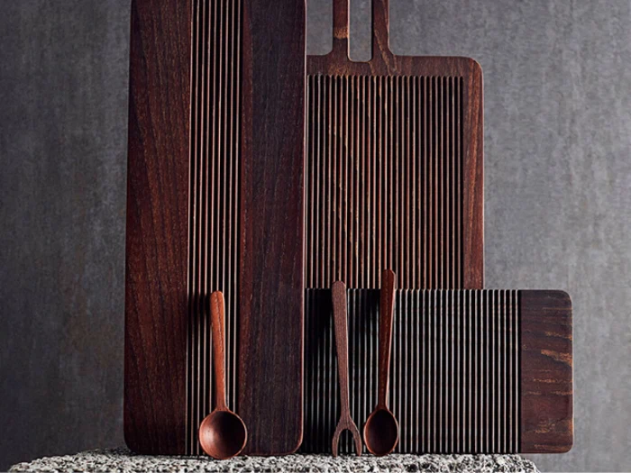 Tre mørke skærebrætter af træ med lodrette riller, en ske og to gafler er anbragt lodret mod en grå struktureret baggrund.