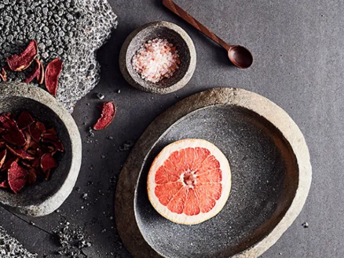 En skive grapefrugt på en stentallerken, med skåle med rosa salt og tørret frugt i nærheden, alt sammen på en mørk, struktureret overflade.
