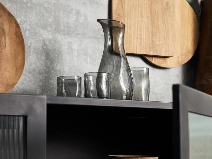 En karaffel af klart glas og tre matchende glas står på et mørkt skab, og bag dem hænger skærebrætter af træ på en grå væg.