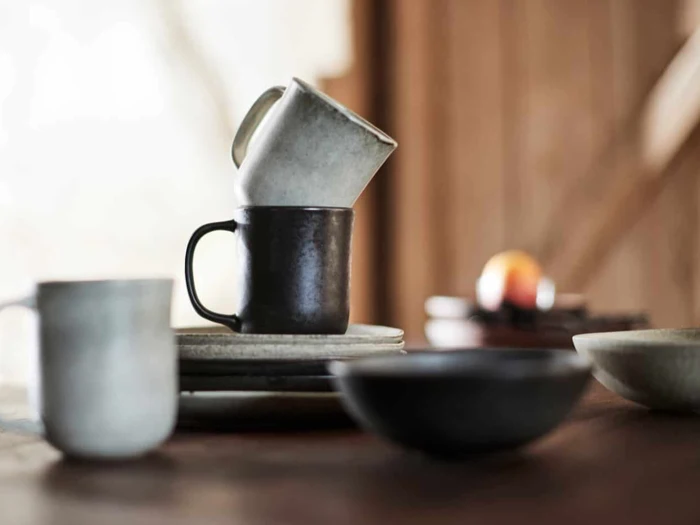 Et nærbillede af keramikkrus stablet på tallerkener med skåle i nærheden på et træbord i et svagt oplyst rum. Baggrunden er sløret.