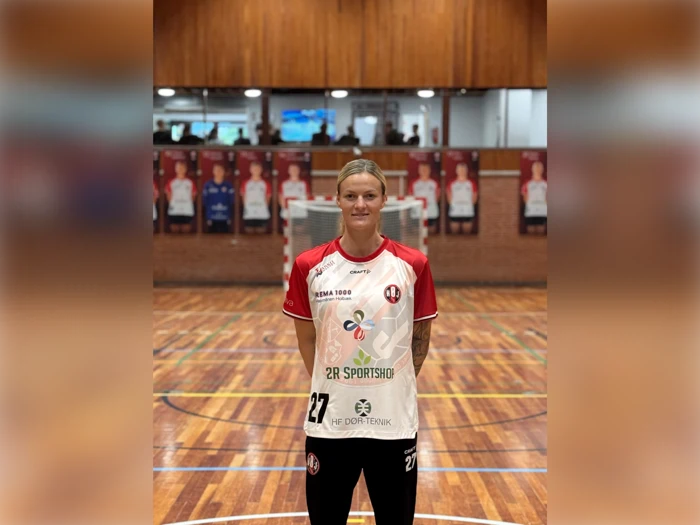 En kvindelig håndboldspiller står på en træbane iført en hvid og rød holdtrøje med sponsorlogoer og nummer 27.