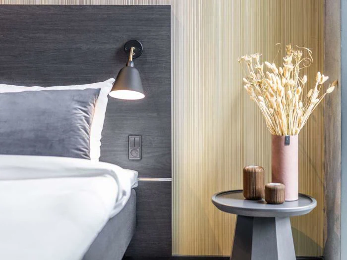 Et moderne soveværelse med en grå seng, hvidt sengetøj, en sort lampe på væggen og et rundt sidebord med en lyserød vase med tørrede planter og to små træbeholdere.