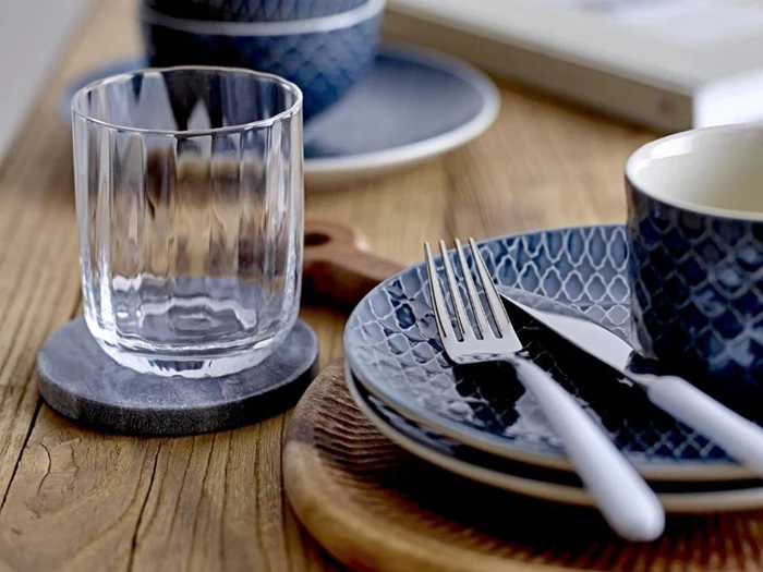 Et klart glas på en bordskåner ved siden af blåmønstrede tallerkener, en kop og gaffel og kniv med hvidt skaft på et træbord.