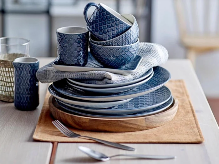 En stak blåmønstrede tallerkener, skåle og krus står på en træbakke med et sammenfoldet håndklæde ved siden af bestik på et bord.