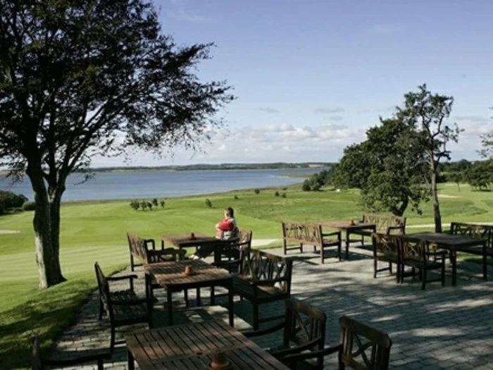 Træborde og -stole på en terrasse med udsigt over en grøn golfbane og en rolig sø, hvor en person sidder og nyder den naturskønne udsigt under en klar, blå himmel.