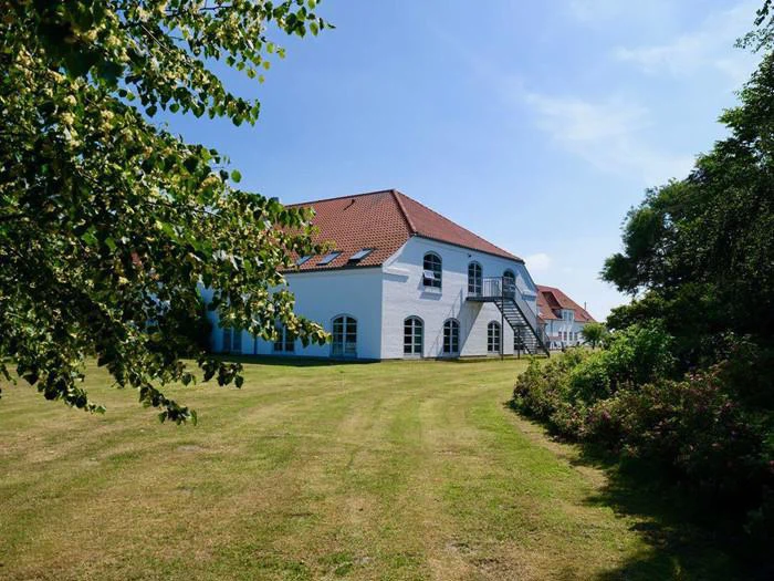 Et stort hvidt hus med rødt tegltag står på en velholdt græsplæne, omgivet af grønne træer og buske under en klar blå himmel.
