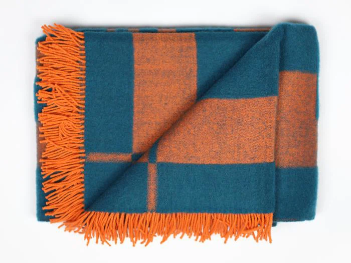 Et foldet uldtæppe med et blågrønt og orange ternet mønster og orange frynsede kanter, lagt på en hvid baggrund.