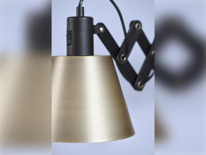 Nærbillede af en moderne messingfarvet lampe med en justerbar sort harmonika-lignende arm på en lyseblå baggrund.