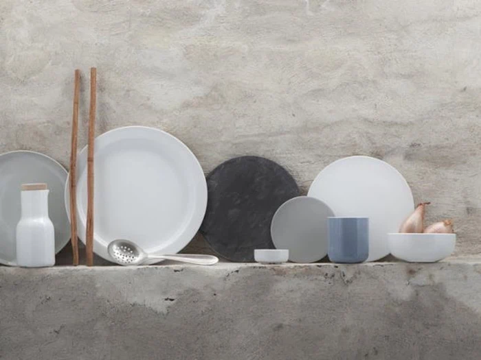 Minimalistisk opstilling af hvide og grå keramiktallerkener, skåle, kopper, en karaffel og træredskaber mod en struktureret, lys væg.