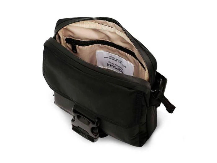 En lille sort taske med lynlås og beige interiør, spænde foran, indvendige lommer og et synligt mærke på indersiden.