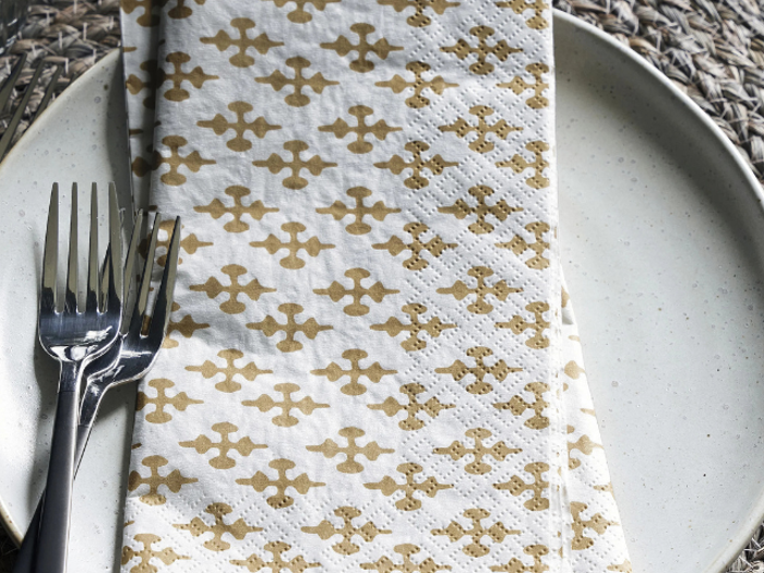 En mønstret serviet med et geometrisk design i guld ligger på en hvid tallerken med to gafler ved siden af.