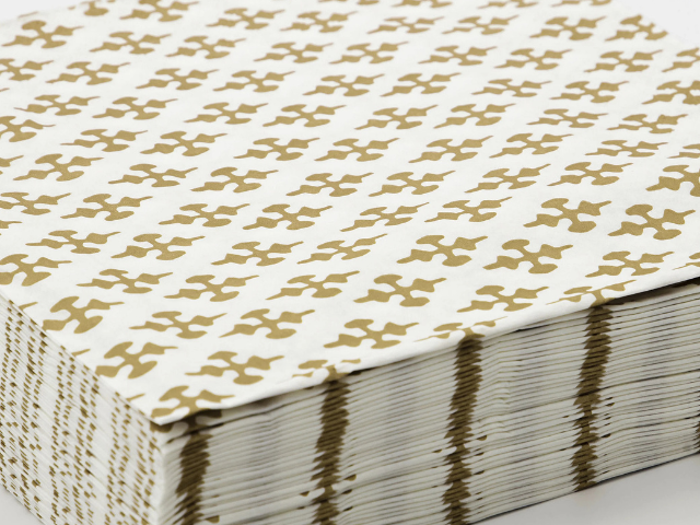 Et nærbillede af en stak hvide servietter med et gentagende geometrisk mønster i guld.
