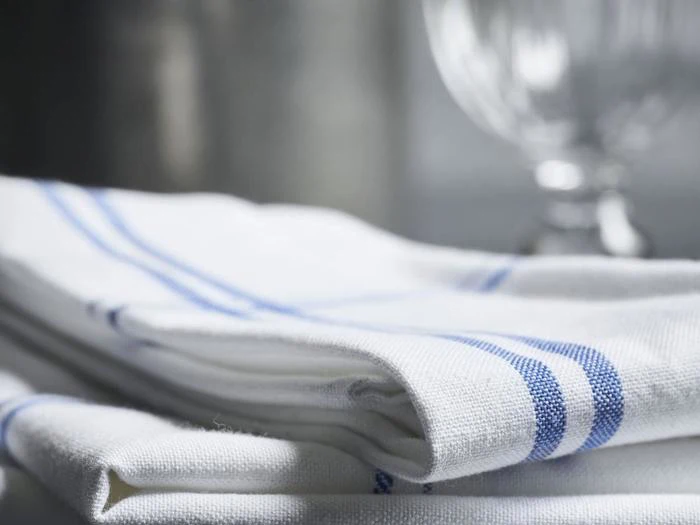Et nærbillede af foldede hvide viskestykker med blå striber, placeret på en overflade. Et klart glas med lodrette riller er sløret i baggrunden.