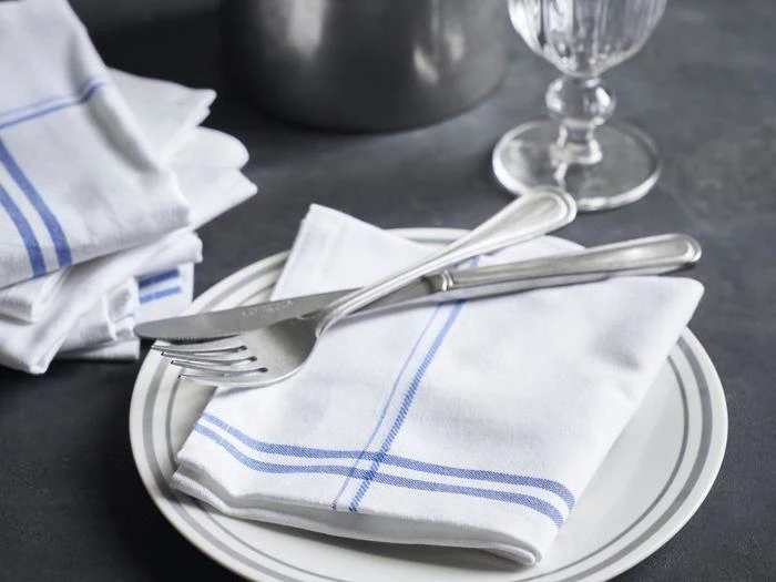 En hvid tallerken med en foldet hvid serviet med blå striber, toppet med en gaffel og kniv, ved siden af en stak servietter, et glas og en metalkande på en mørk overflade.