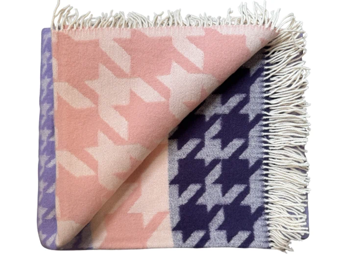 Et blødt tæppe med et stort hundetandsmønster i pink, lilla og cremefarver, med frynser langs den ene kant og et pænt foldet hjørne.