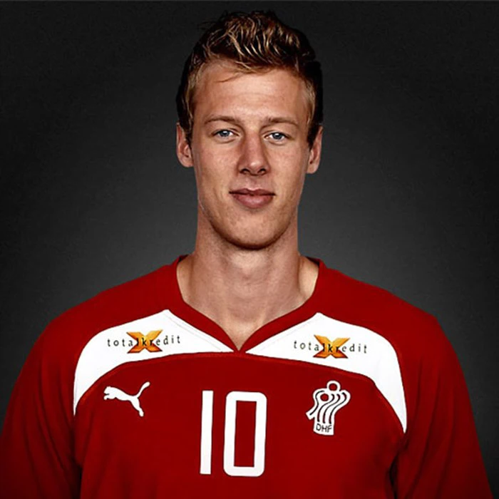 En mand i en rød sportstrøje med nummer 10 og hvide logoer står på en mørk baggrund og kigger ind i kameraet.