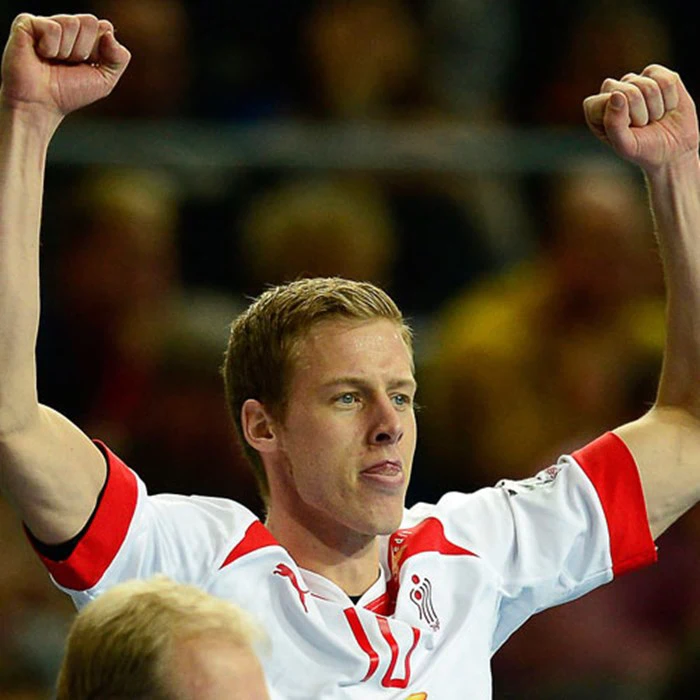 En mandlig atlet i en hvid sportstrøje med røde detaljer løfter begge arme i jubel med et fokuseret udtryk i ansigtet.