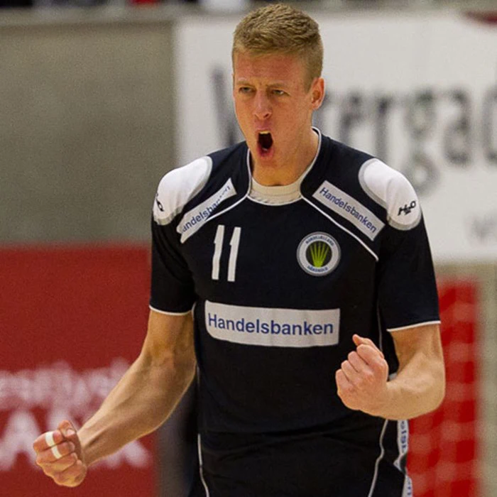 En mandlig håndboldspiller i en mørk trøje jubler med knyttede næver og åben mund efter et spil og viser intense følelser.