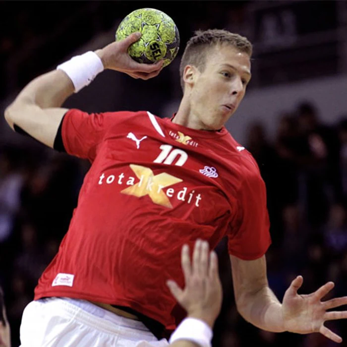 En mandlig håndboldspiller i rød trøje hopper og gør sig klar til at kaste en grøn bold, mens en anden spillers hånd er synlig i forgrunden.