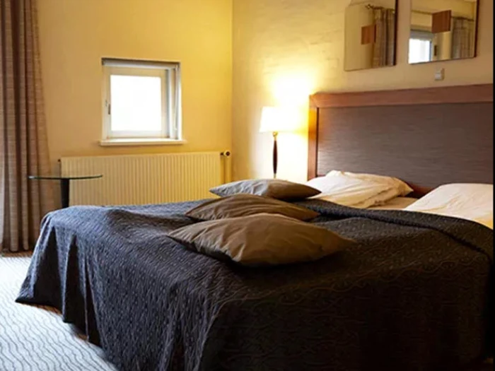 Et hyggeligt soveværelse med en dobbeltseng, mørkt sengetæppe og to brune puder. En lampe, et lille vindue og vægkunst er synlige og skaber en varm og indbydende atmosfære.