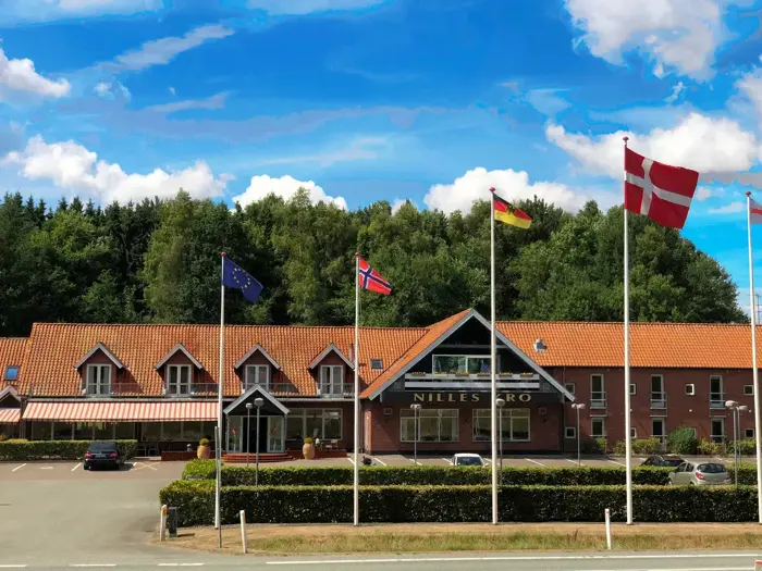 En murstensbygning med rødt tegltag, omgivet af træer. Fire flag, deriblandt Danmarks, vajer foran under en blå himmel med skyer.