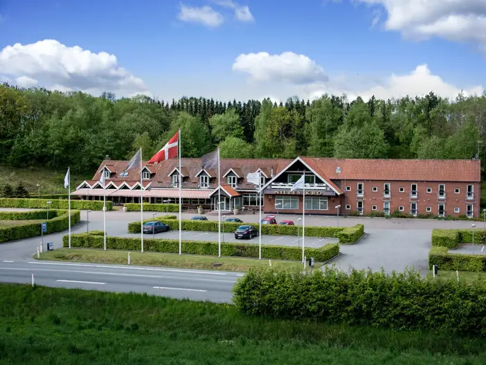 En stor murstensbygning med rødt tegltag, danske flag og en parkeringsplads med biler, omgivet af grønne områder og træer under en delvis overskyet himmel.