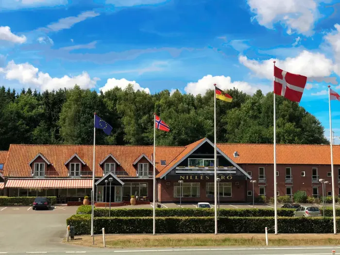 Et hotel med rødt tegltag, flere flag foran og træer i baggrunden under en blå himmel med skyer.
