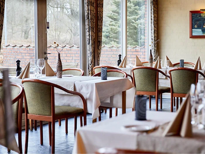 En lys restaurant med hvide duge, pænt opstillede borde, træstole med grønt betræk og store vinduer ud til en have.