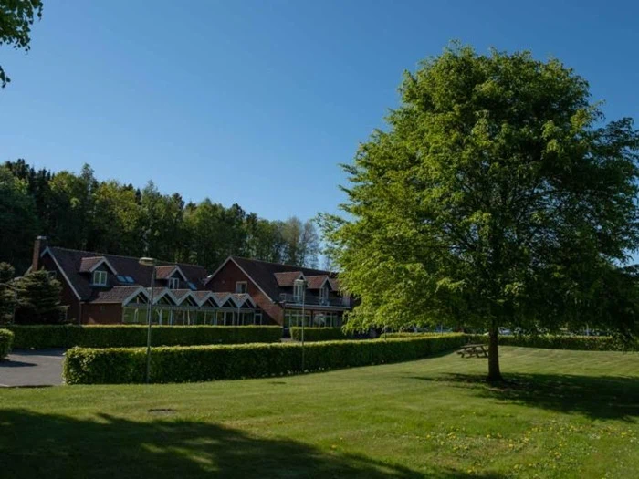 Et stort murstenshus med rødt tag ligger bag en sirligt klippet hæk ved siden af en frodig grøn græsplæne og et stort løvtræ under en klar blå himmel.