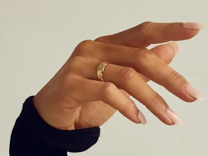 En hånd med lange, velplejede negle bærer en guldring med struktur på langfingeren, set mod en ensfarvet baggrund. Personen er iført en sort ærme.