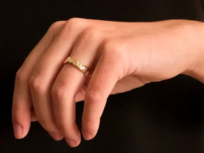 En hånd med en ring i guldtekstur på ringfingeren, fotograferet på en ensfarvet sort baggrund.