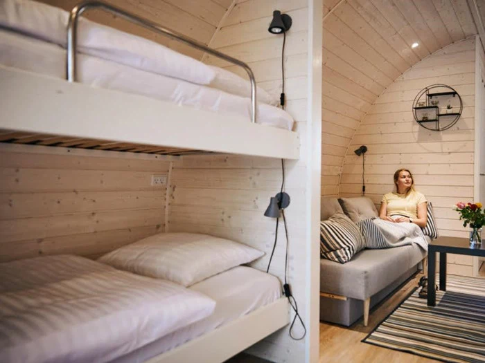 En kvinde sidder i en sofa i et hyggeligt træhytterum med køjesenge, stribede puder og et lille bord med blomster.