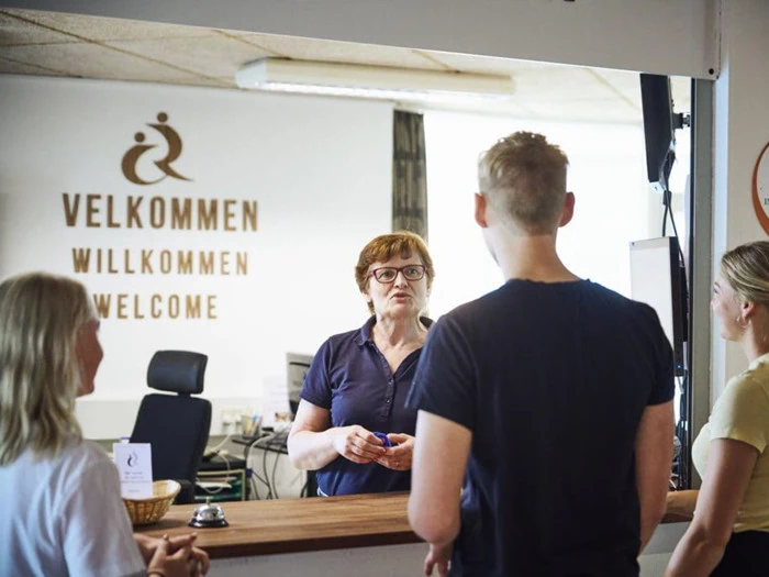 En kvinde står bag en receptionsskranke og taler med tre personer; på et skilt bag hende står der Velkommen på flere sprog.