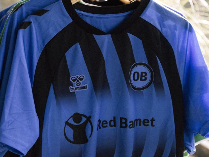 En blå- og sortstribet fodboldtrøje med Hummel-logo, OB-våbenskjold og Red Barnet-sponsor på forsiden, hængende på et stativ.