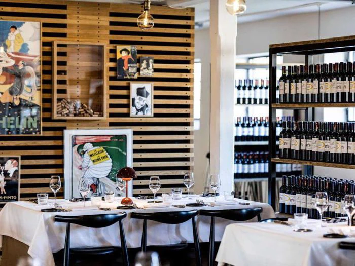 Et moderne restaurantinteriør med hvide duge, dækkede borde, sorte stole, vægkunst og hylder fyldt med vinflasker.