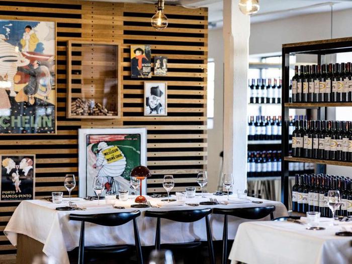 Et moderne restaurantinteriør med dækkede borde, vinflasker på hylder og vintage Michelin-plakater på væggen.
